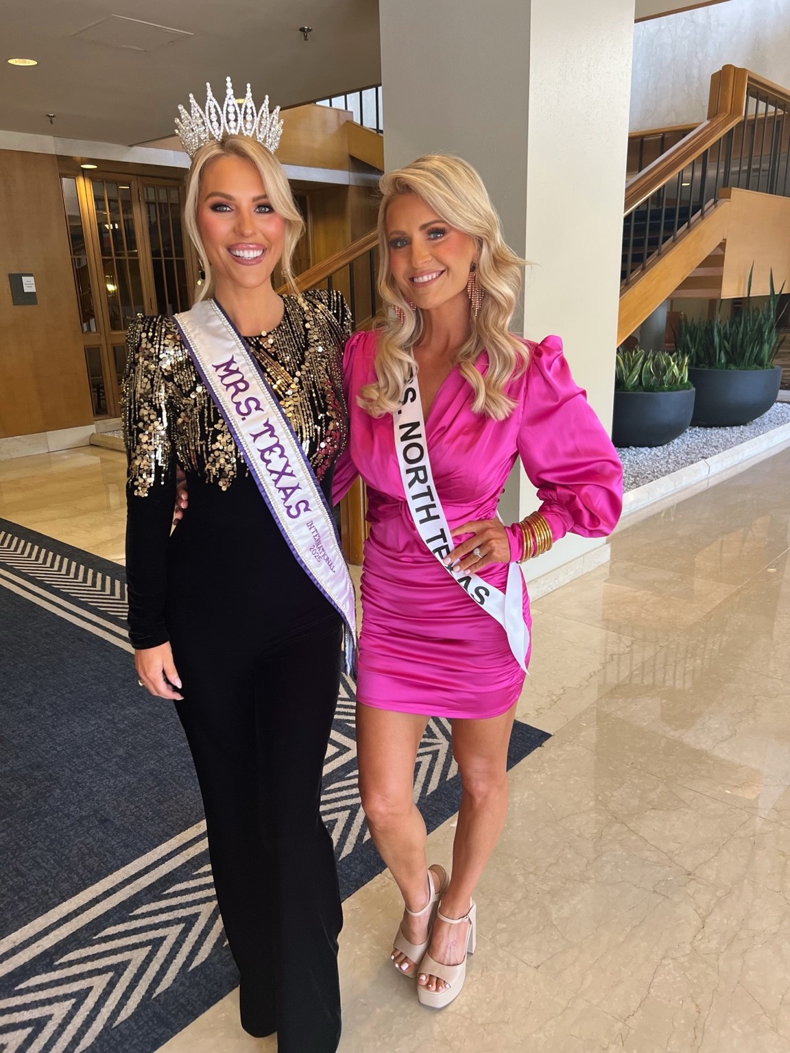 Two pageant titleholders smiling together in a lobby, wearing black and pink